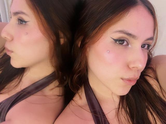 Photographie en gros plan d'AmberlStha avec de longs cheveux bruns, portant un haut noir à encolure américaine, reflétée dans une prise de vue à double face. Elle a un trait d'eye-liner noir épais, et une petite tache de beauté sur sa joue. Incroyablement séduisant, regard direct..