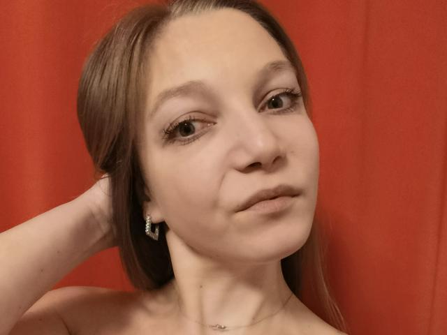 Nahaufnahme von LovelyxEmilly, einer hellhäutigen Frau mit braunen Haaren, die Hand hinter dem Kopf hat, roter Hintergrund, neutraler Gesichtsausdruck. dezent geschminkt, kleine Ohrringe, Halskette, leichte Rouge, leichte sexuelle Anspannung, lässige Pose.