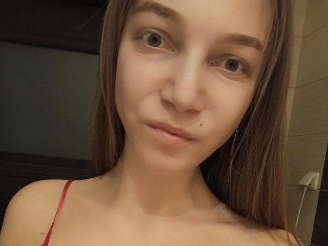 Close-up de LovelyxEmilly, uma jovem com pele clara, olhos castanhos e cabelos longos castanhos, vestindo uma blusa vermelha com alças. Ela tem uma pequena marca acima do lábio superior. Incrivelmente fofa, com uma expressão ligeiramente nervosa..