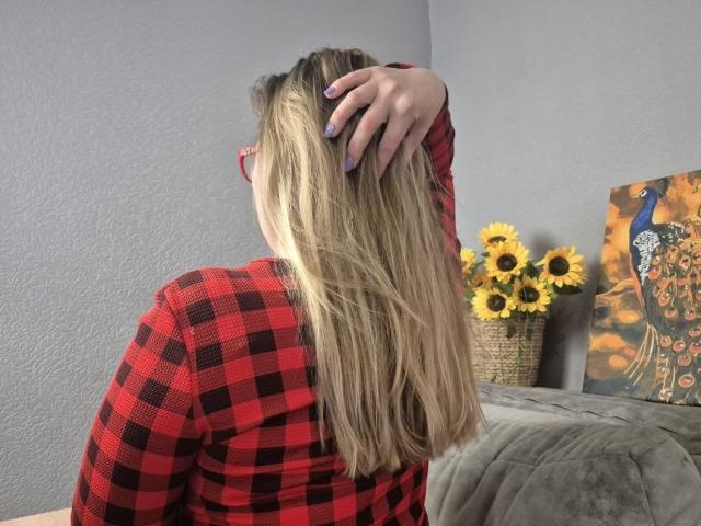 Viesta, de espaldas, cabello rubio, camisa de cuadros rojos, mano en el cabello, uñas pintadas de morado. Girasoles en una cesta, pintura colorida, sofá gris. Tensión sexual, ambiente provocador, informal, pose relajada..