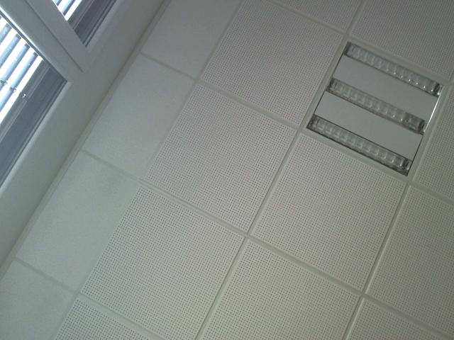 Gros plan d'un plafond avec des panneaux perforés et une grille d'aération rectangulaire. L'ombre en forme de pénis de Olegator-hot se projette sur la droite. Une lumière vive provenant d'une fenêtre se trouve sur la gauche. Teinte grisâtre.