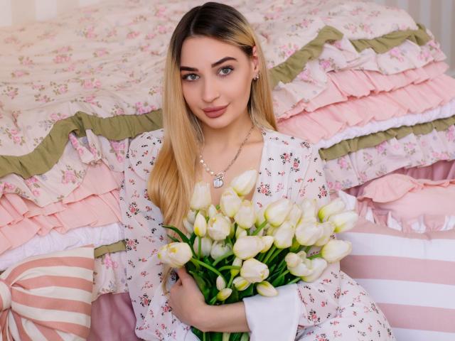 Barbara Rosy mit langen blonden Haaren, die ein weißes Kleid mit Blumenmuster trägt und einen Strauß weißer Tulpen hält, sitzt auf einem Bett. mit rosa, gerüschter Bettwäsche, und direkt in die Kamera blickend. Verdammt heiß..