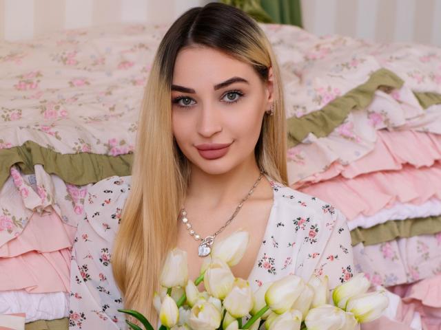 BarbaraRosy with long blonde hair, blue eyes, and pink lips, holding white tulips, wearing a white floral top, sitting in front of a pink and green ruffled bedspread. Fucking sexy.
