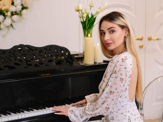 Barbara Rosy, aux longs cheveux, à la peau claire, portant une blouse blanche à fleurs, assise devant un piano noir, regardant en arrière avec un regard séduisant.
fleurs blanches dans un vase sur le piano, éclairage intérieur lumineux..
