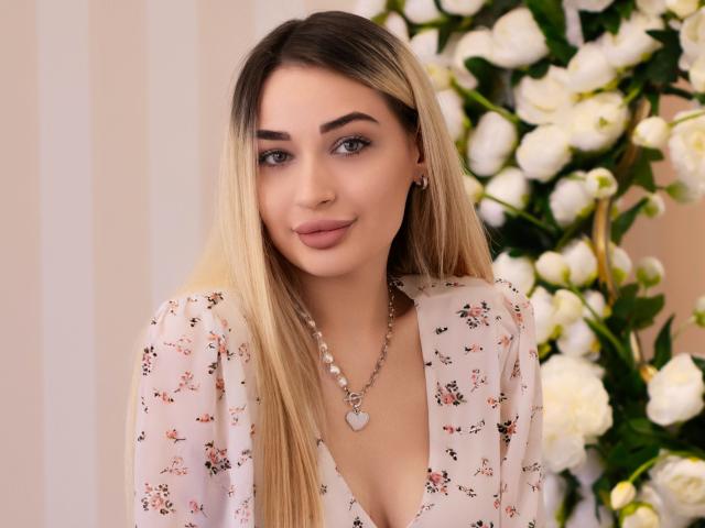 Fotografia de BarbaraRosy, uma mulher com cabelos longos e loiros, vestindo uma blusa branca com estampas florais e um decote em V profundo, e um colar de pérolas. colar, em frente a flores brancas. Muito atraente..