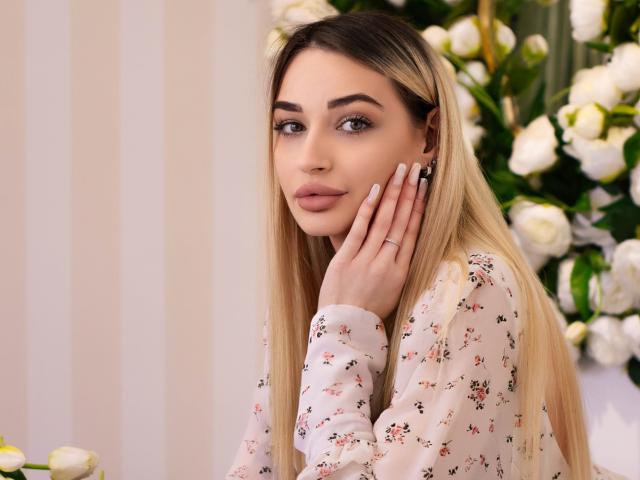 Barbara Rosy, lang blond haar, draagt een witte blouse met bloemen, hand op haar wang, blauwe ogen, zachte uitdrukking, witte bloemen in achtergrond, wazig, zacht licht, seksuele spanning, subtiele aantrekkingskracht, vrouwelijk, aantrekkelijk.