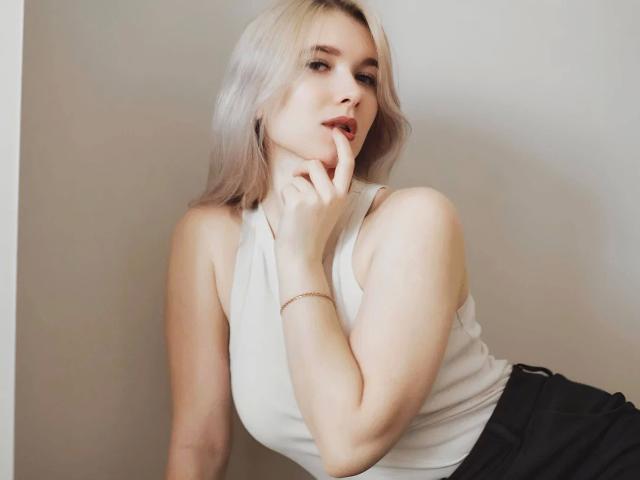Photographie de KarolinaRay-hot, appuyée contre un mur blanc, portant un débardeur blanc et un pantalon noir, avec des cheveux blonds. et un doigt dans sa bouche, dégageant une tension sexuelle et un charme irrésistible..