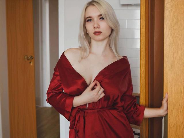 KarolinaRay, com seus cabelos loiros platinados, está em um corredor, vestindo um vestido de cetim vermelho escuro, com um decote que deixa os ombros à mostra, e segurando... o tecido próximo à sua região do colo. Parede branca com azulejos ao fundo. Tensão sexual.