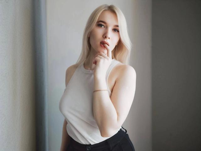 Fotografía de KarolinaRay-hot, una mujer rubia de piel clara, que lleva una camiseta blanca sin mangas y pantalones negros de cintura alta. y una pulsera de oro. Ella está mordiéndose el dedo de forma seductora, con una iluminación suave..