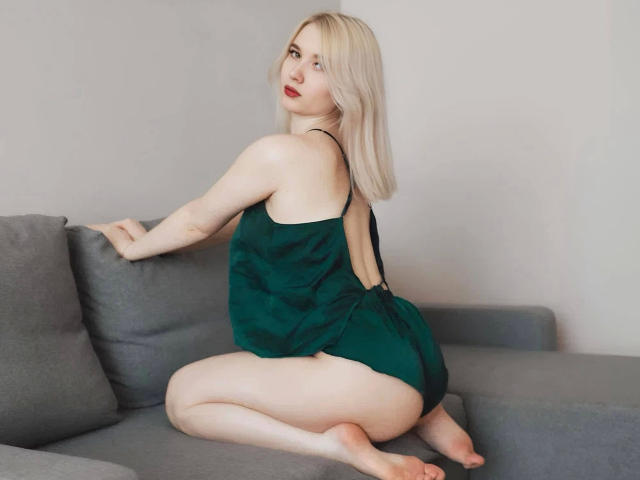 KarolinaRay-hot, una donna bionda dalla pelle chiara e con rossetto rosso, è inginocchiata su un divano grigio, indossando una top nera senza schienale. le gambe divaricate, il pene visibile, sguardo seducente, illuminazione intensa.