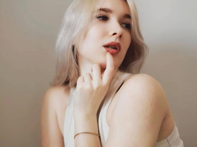 Close-up foto van KarolinaRay, met een bleke huid en platinablonde haren, die verleidelijk aan haar vinger likt. Ze draagt een witte top zonder bandjes, met een gouden armband om haar rechterpols. Heel aantrekkelijk..