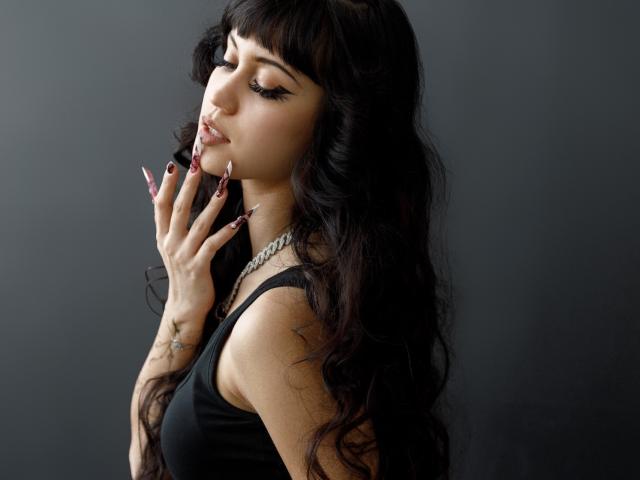 TiffanySweetex, een donkerharige vrouw met lang, golvend haar, een zwarte tanktop, een zilveren ketting, met haar hand in de buurt van haar gezicht, kijkend. Verleidelijk, donkere achtergrond, scherpe belichting, tatoeage op de hand, donker geverfde nagels. Heel aantrekkelijk..