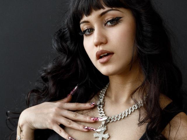 TiffanySweetex a les cheveux noirs et ondulés, un trait d'eye-liner noir épais et des lèvres pulpeuses. Elle touche sa poitrine avec ses ongles soignés, tout en portant un collier en argent avec un grand pendentif en forme de papillon. Fond noir.