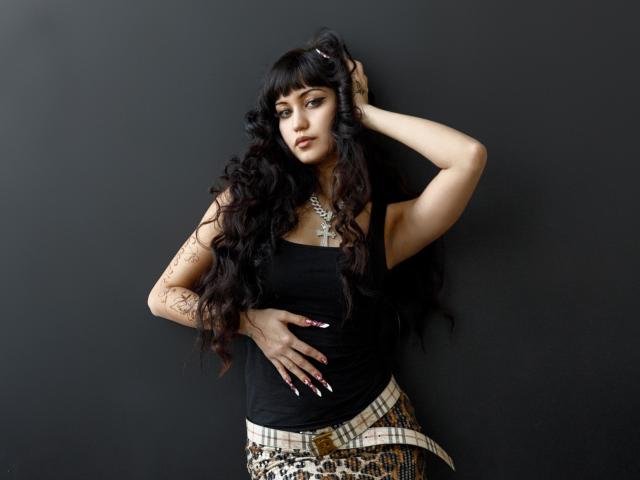 TiffanySweetex, eine kurvige Frau mit langen, schwarzen Haaren, lehnt sich an eine schwarze Wand. Sie trägt ein schwarzes Oberteil, einen karierten Rock und eine Sternkette, ihre Hand liegt auf ihrem Bauch, und sie strahlt eine verführerische Spannung aus..