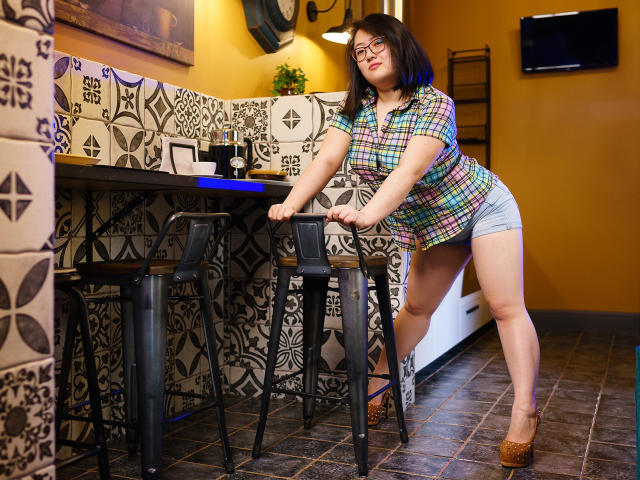 QiraNiki, curvy with large ass, wearing a colorful plaid shirt and blue shorts, leans on black bar stools, her hands on the metal, in a tiled room.