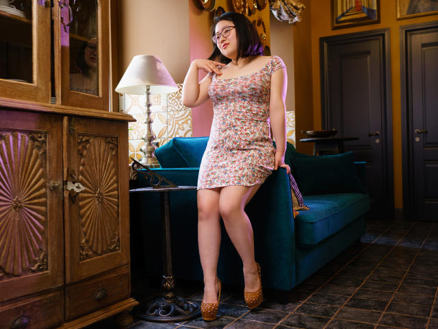 QiraNiki, wearing a floral dress, stands in a room with a blue couch, wooden cabinet, and lamp. She has purple-tipped black hair, glasses, and brown high heels. Her tits are visible.