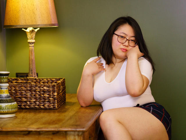 QiraNiki with big tits, glasses, and black hair, sitting on a wooden table, hands on face, wearing a white top and plaid skirt, next to a wicker lamp.