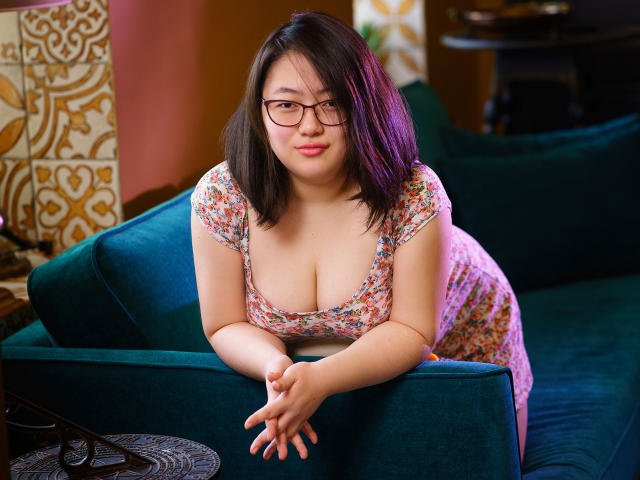 QiraNiki with medium brown skin, black glasses, and pink lipstick leans on a green couch. She has purple-tipped black hair, floral dress, and shows her big, round tits.