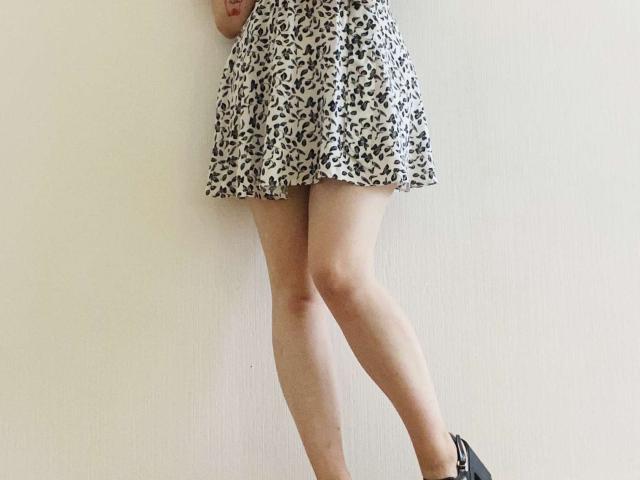 KarKutie's legs and lower body, wearing a short, black-and-white floral skirt, standing against a white wall, with a black heel on her right foot. Fucking sexy, skirt barely covers her cunt.