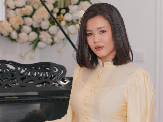 ArianaFlower, con i capelli neri lisci, indossa una camicia gialla con bottoni ed è in piedi di fronte a un pianoforte nero decorato. Rose bianche sullo sfondo. Lei guarda direttamente la telecamera, con un leggero sorriso..