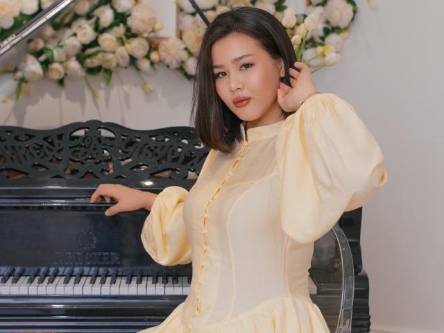 ArianaFlower, avec une peau de couleur moyenne et des cheveux noirs mi-longs, portant une robe jaune à manches longues, se tient debout. Devant un piano noir, avec des fleurs blanches en arrière-plan. Elle touche ses cheveux..