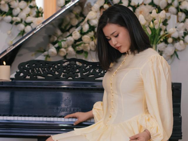 ArianaFlower, con cabello oscuro, vestida con un vestido amarillo con botones, está sentada frente a un piano negro, tocando con concentración. expresión, rodeada de flores blancas. La luz suave resalta su figura curvilínea..