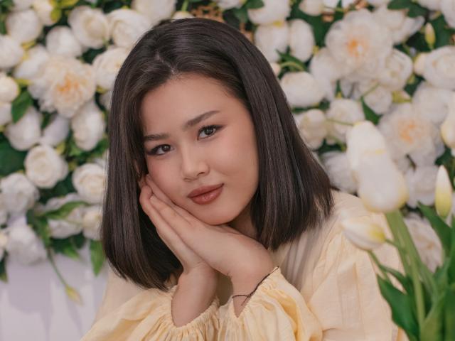 ArianaFlower with straight black hair, pale skin, and red lipstick, resting her chin on her hand, wearing a yellow blouse, surrounded by white roses. Sexy, flirty expression, soft lighting.