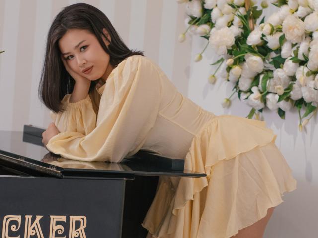 ArianaFlower, eine asiatische Frau mit schulterlangem, schwarzem Haar, beugt sich über ein schwarzes Klavier und trägt ein gelbes Kleid mit Rüschen. Weiße Blumen im Hintergrund. Eine unglaublich sexy Pose. CAFR-Logo unten links..