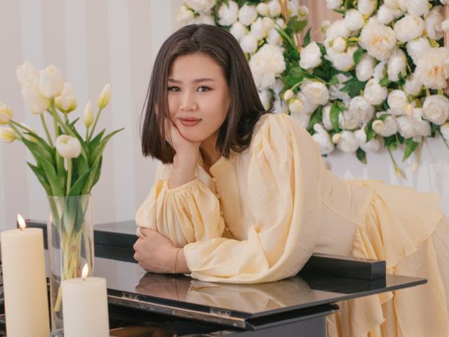 ArianaFlower mit schulterlangem, schwarzem Haar, die an einem schwarzen Tisch lehnt und ein gelbes Kleid trägt. Weiße Kerzen, weiße Tulpen und blühende Blumen im Hintergrund. Verdammt sexy, entspannter Gesichtsausdruck..