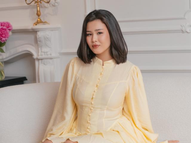 ArianaFlower, uma mulher asiática com cabelo preto na altura dos ombros, está sentada em um sofá branco, vestindo um vestido amarelo com botões. Ela está olhando para a câmera com um sorriso discreto, transmitindo uma aura sexy e confiante..