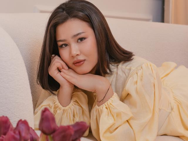 ArianaFlower with short black hair, leaning on a white couch, wearing a yellow blouse, resting her head on her hand, looking at the camera, with red rose petals in the foreground.