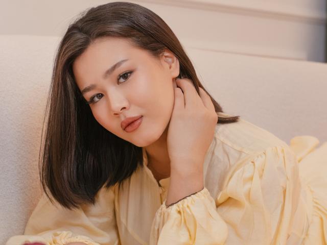 ArianaFlower mit glattem, schwarzem Haar, die ein gelbes Hemd trägt, lehnt sich an ein weißes Sofa, ihre Hand berührt ihren Hals, ihr Ausdruck ist ernst. Ausdruck, weiches Licht, dezentes Make-up, sexuelle Spannung, Nahaufnahme.