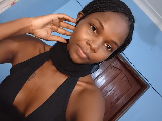 Photograph of PrincesLove, an attractive black woman with braided hair, wearing a black halter top, exposing her cleavage and small nipple tattoo. She's smiling, touching her hair, with a wooden door in the background.