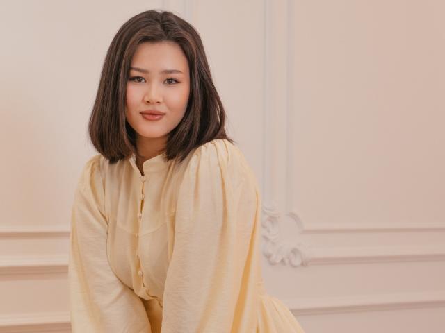 ArianaFlower, avec ses cheveux noirs mi-longs, portant une chemise blanche cassée ample et ouverte, penchée légèrement en avant, sourire doux, tension sexuelle subtile, mur blanc éclatant avec moulures décoratives en arrière-plan.