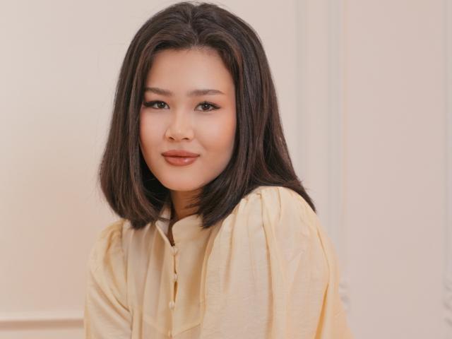 Fotografia di ArianaFlower con capelli neri di media lunghezza, che indossa una camicia gialla con bottoni, e guarda direttamente verso... con una fotocamera e un sorriso appena accennato. Pelle chiara, leggero trucco, illuminazione soffusa, leggera tensione sessuale.