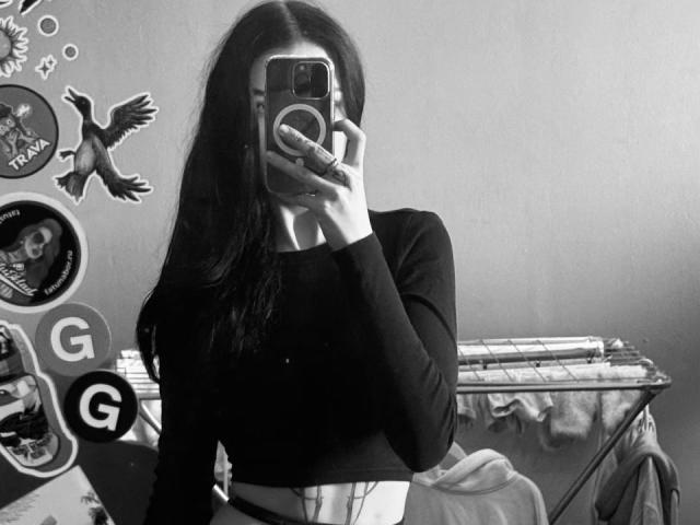 Black and white photo of ShylaCatillo with long dark hair, wearing a black long-sleeve top, taking a mirror selfie with her phone, covering her face. Stickers, bird, and clothes in background.