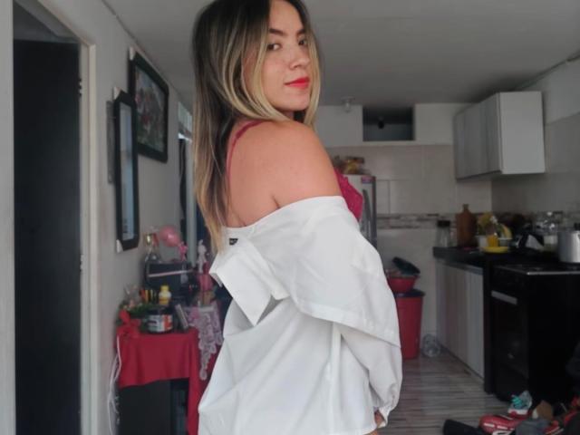 SexySussieX com cabelos castanhos longos, batom vermelho e uma camisa branca com um ombro de fora, olhando para trás com um sorriso sedutor. Contexto: cozinha com mesa vermelha, armários brancos e objetos espalhados..