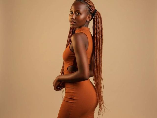 Sexyariaashley-hot se tient de côté, regardant par-dessus son épaule. Elle porte une robe orange moulante qui met en valeur ses courbes et ses longs cheveux tressés. Sa peau foncée contraste avec le fond chaleureux. Incroyablement sexy..