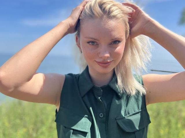 Close-up photo of CrystalHope, blonde woman with fair skin, blue eyes, and small breasts, wearing a green sleeveless shirt, hands in blonde hair, bright daylight, blue sky, green field background.