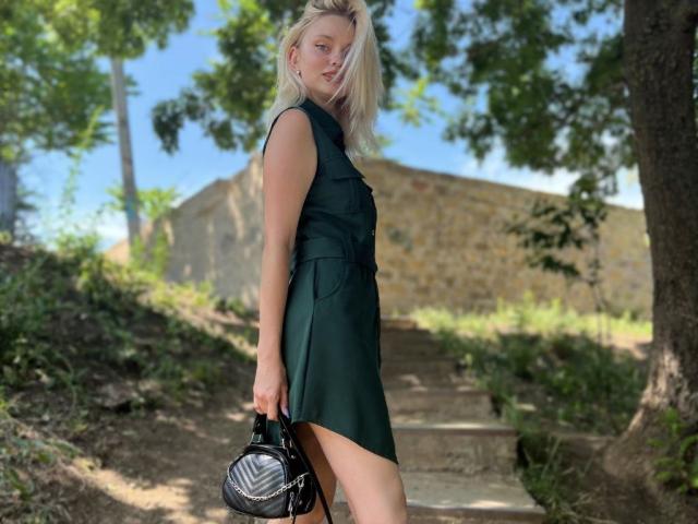 CrystalHope, eine blonde Frau, trägt ein schwarzes, ärmelloses Kleid und hält eine schwarze Handtasche. Sie steht auf steinernen Stufen, im Hintergrund sind Bäume und ein blauer Himmel zu sehen. Hintergrund, sexy, schlank, attraktiv, Sonnenlicht betont ihre Figur, Außenaufnahme..