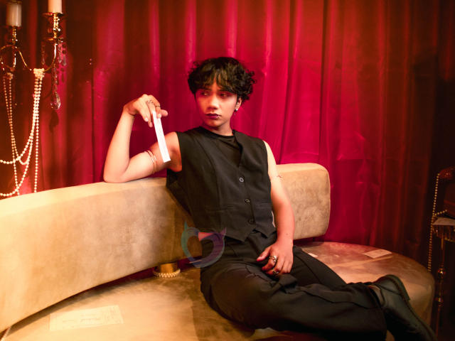 EriolWest with messy black hair, wearing a black vest and pants, sits on a beige couch, holding a white feather. Red curtains and string lights create a sultry, seductive atmosphere.