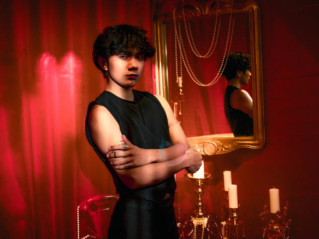 EriolWest, standing with arms crossed, wearing a black sleeveless shirt, in a dimly lit room with red curtains. Reflection in ornate mirror. Candles on a table, sexy vibe.