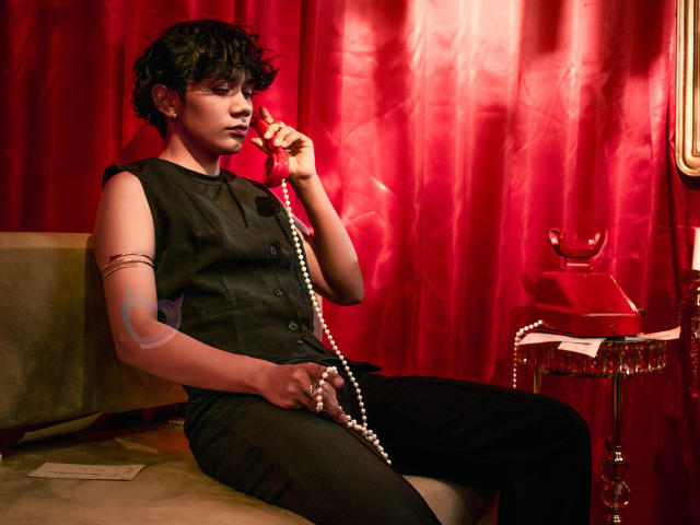 EriolWest, avec ses courts cheveux noirs, est assise sur un canapé, tenant un téléphone avec un cordon de perles, et portant une robe noire sans manches. Des rideaux rouges, une table en verre avec un bol. Putain, c'est sexy..