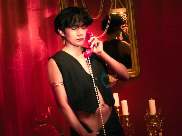 EriolWest, avec des cheveux noirs courts, portant un haut noir sans manches, tenant un téléphone rouge à l'oreille, un collier de perles autour du cou. Au-dessus de la poitrine, éclairage rouge, deux bougies en arrière-plan, pose sexy et séduisante..