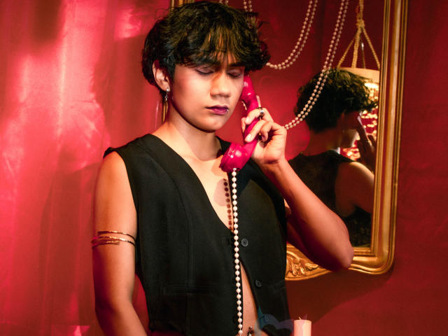 EriolWest, kurze, dunkle Haare, schwarzes Top ohne Ärmel, hält ein rosa Telefon ans Ohr, rotes Licht, Spiegelung in einem goldenen Spiegel, Perle. Halskette, Armband, sinnlicher Ausdruck, sexuelle Spannung, roter Hintergrund..