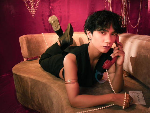 EriolWest, allongée sur un canapé beige, les jambes en l'air, vêtue de noir, tenant un téléphone, des perles enroulées autour du cou, fond rouge, chaussure dorée étincelante, regard intense, sexy, pose séduisante, style décontracté.