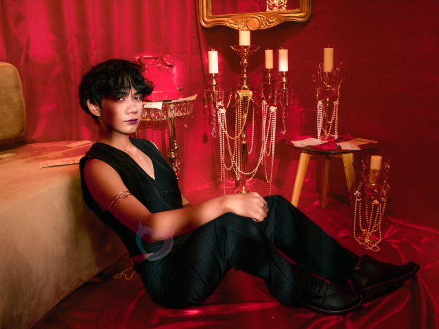 EriolWest with short black hair sits on red satin, wearing a black sleeveless top and pants, surrounded by glowing candles and beaded ornaments. Red light casts sultry shadows. Fucking sexy.