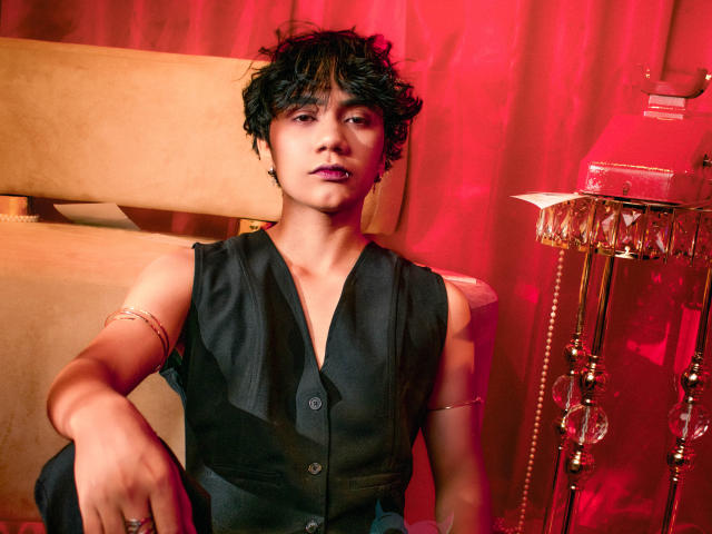 EriolWest, met kort, rommelig zwart haar, gekleed in een zwart vest zonder mouwen, zittend in een kamer met rood licht. Arm op de stoel, rode gordijnen, lamp met franjes. Sensuele, zelfverzekerde pose..