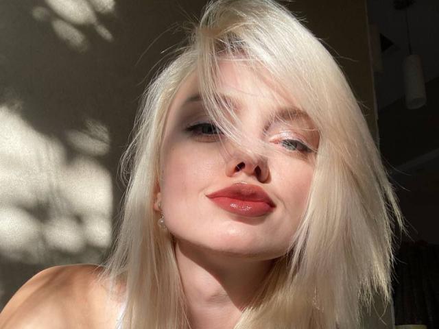 Close-up foto van CrystalHope met platinablond haar, een door de zon verlicht gezicht en rode lippen. Haar haar bedekt gedeeltelijk haar ogen, waardoor er schaduwen ontstaan. Helder zonlicht accentueert haar lichte huid. Heel erg aantrekkelijk..