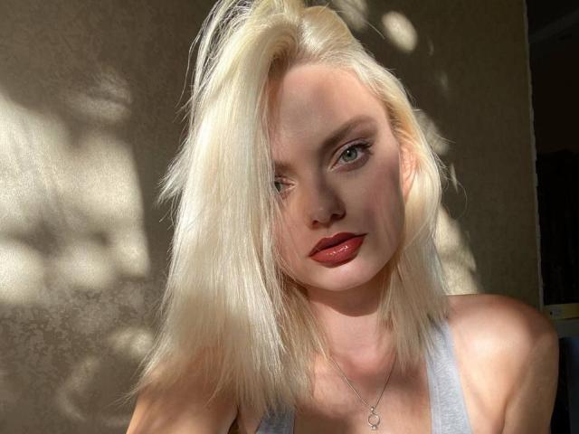 Fotografia de CrystalHope com cabelo loiro platinado, olhos azuis intensos e batom vermelho, vestindo uma camiseta branca. Iluminado pela luz do sol, com sombras projetadas em uma parede texturizada. Incrívelmente atraente, olhar direto..